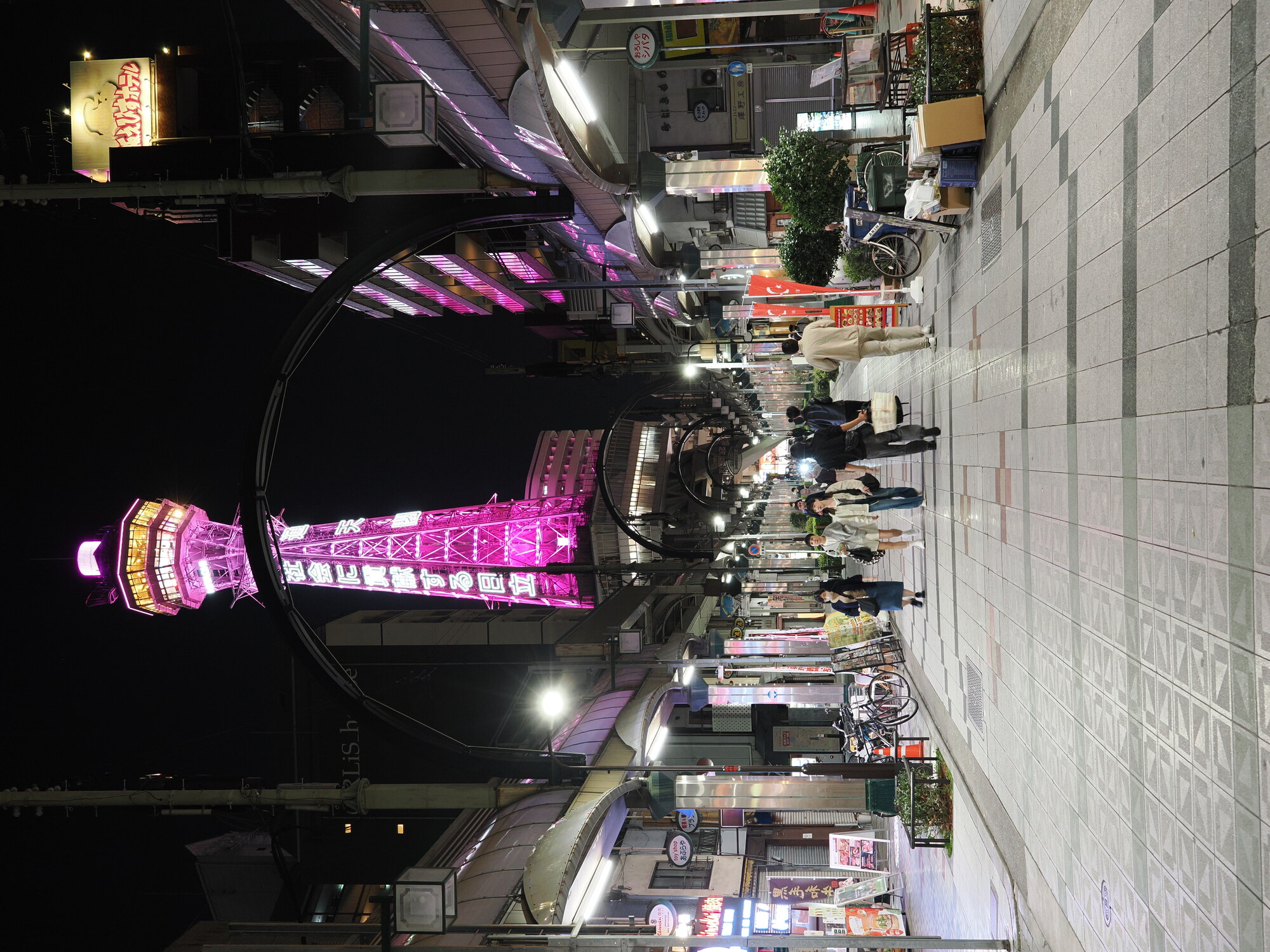 Tsutenkaku nocą — wieża Tsutenkaku w różowo-magentowym oświetleniu przy końcu alei Shinsekai, reklama Hitachi 社会に貢献する日立, napis 通天閣 na łuku nad ulicą