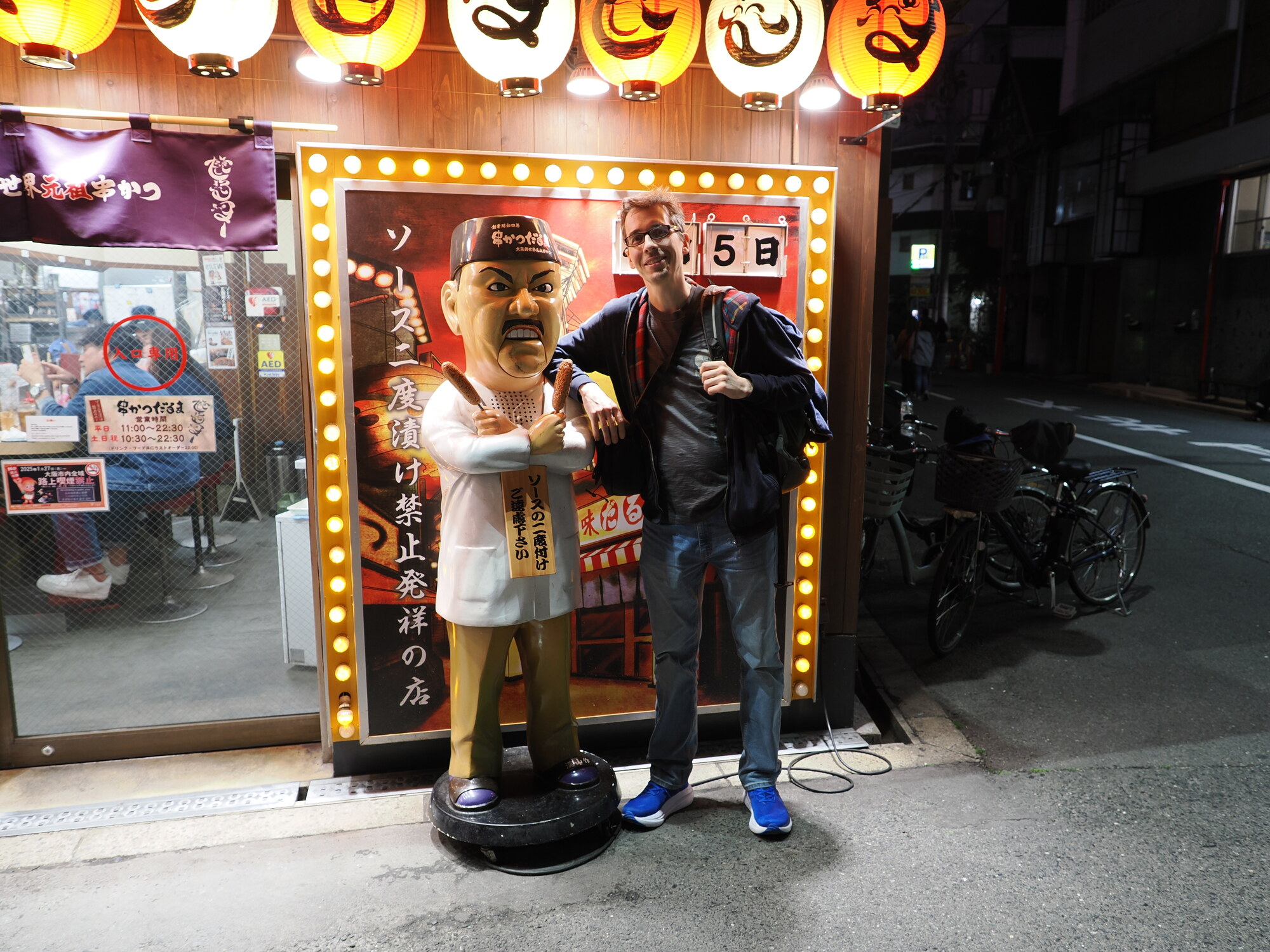 Kamil przy maskotce Daruma restauracji Kushikatsu Daruma w Shinsekai — mężczyzna pozuje przy figurce właściciela z szaszłykami, tabliczka ソースに二度漬け禁止発祥の店