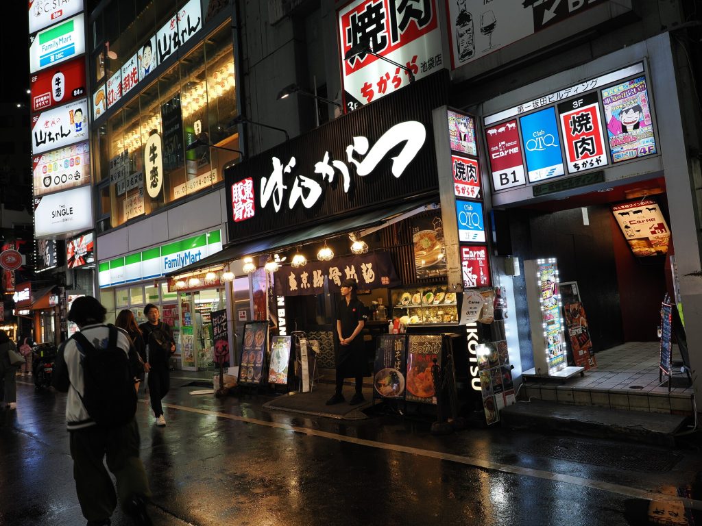 Restauracja ばんかつ Bankatu ramen nocą w Ikebukuro - czarny szyld z białymi kanji, obsługa przy wejściu, FamilyMart obok, mokra ulica