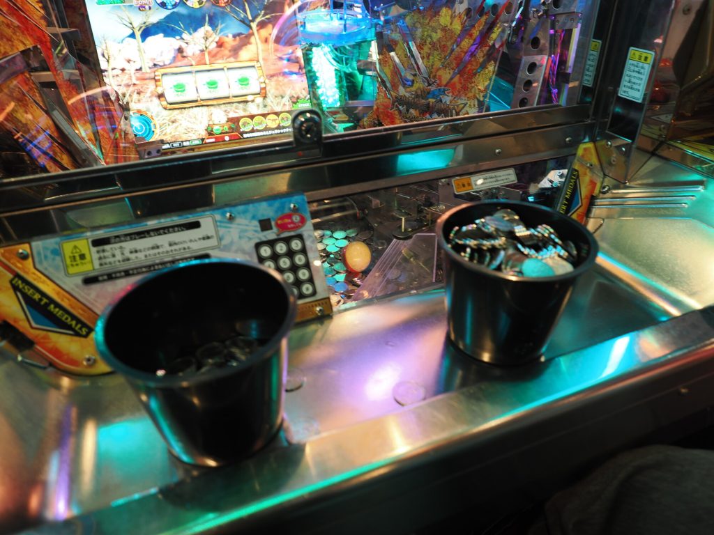 Dwa kubki z żetonami na medal game ROUND1 - INSERT MEDALS, niebieskopurpurowe oświetlenie, ekran gry fantasy w tle