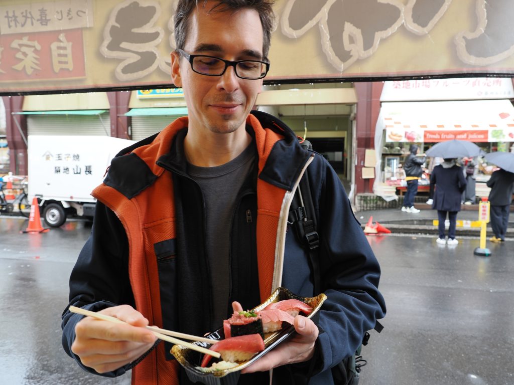 Kamil z pałeczkami je nigiri z tuńczykiem stojąc na ulicy Tsukiji w deszczu